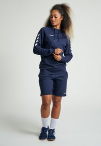 Mörkblå huvtröja med vitt logotyp, dragsnörning och sidokontraster, ihopparad med matchande shorts och vita sportstrumpor.