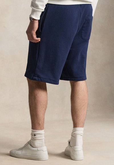 Man wearing navy blue shorts, white crew socks, white sneakers, and a white long-sleeve top, standing against a neutral background.