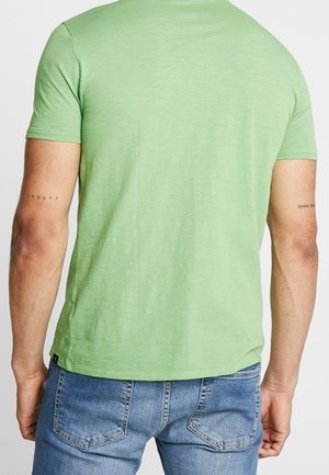 Man die een lichtgroen t-shirt met korte mouwen en blauwe spijkerbroek draagt, van achteren gezien met zichtbare tatoeages op beide armen.