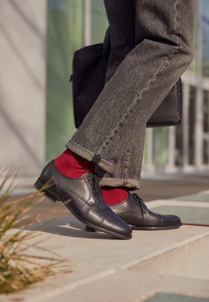 Schwarze Lederschuhe mit Schnürsenkeln, kombiniert mit hochgekrempelten grauen Hosen, roten Socken und einer schwarzen Tasche in einer gut beleuchteten Außenumgebung.