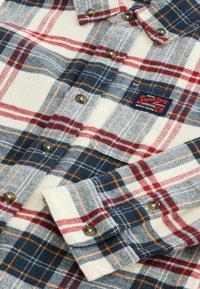 Flannelskjorte med ternet mønster i marineblå, rød og creme, med knappeaccenter og en lille brandmærke tæt på kraven.