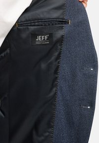 Navy herringbone blazer interieur met zwarte voering, een ritszak en een label met de tekst "JEFF URBAN TAILORING" in het wit.