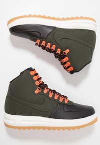 High-top sneakers featuring a dark green synthetic upper, black rubber accents, orange eyelets, and a gum rubber outsole.