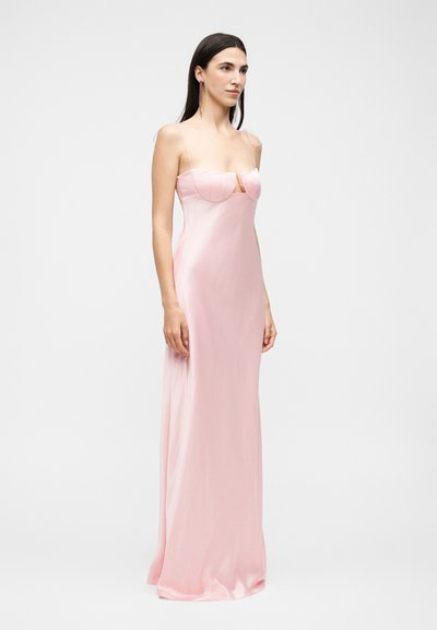 Γυναίκα με μακριά σκούρα μαλλιά, που φορά ένα εφαρμοστό, μακρύ μέχρι το πάτωμα, απαλό ροζ σατέν φόρεμα με λεπτές τιράντες και διαμορφωμένη λεπτομέρεια στο στήθος.