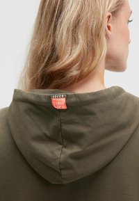 Olivgrüner Hoodie mit einer runden Kapuze und eingenähtem Etikett am Hals. Weiche, glatte Textur mit minimalen Details.