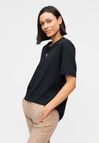 Μαύρο κοντομάνικο μπλουζάκι με στρογγυλή λαιμόκοψη και μικρή λεπτομέρεια λογότυπου στο στήθος, συνδυασμένο με μπεζ παντελόνι που διαθέτει πλαϊνές τσέπες.