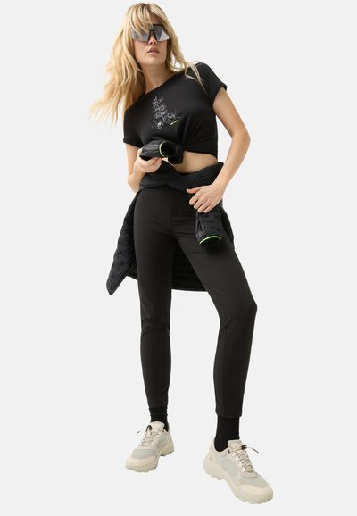 Black cropped T-shirt with graphic design, paired with black slim-fit pants. Model wears a black puffer jacket tied at the waist and chunky white sneakers.