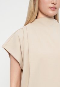 Blusa beige con collo alto e maniche corte, strutturate. La texture liscia e le pieghe sottili aggiungono dimensione al design.