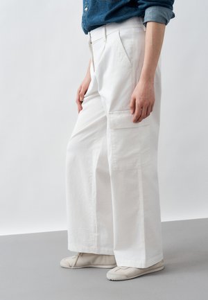 Άτομο που φοράει λευκό παντελόνι cargo με φαρδιά μπατζάκια και ανοιχτόχρωμα sneakers, στέκεται μπροστά σε απλό φωτεινό φόντο.