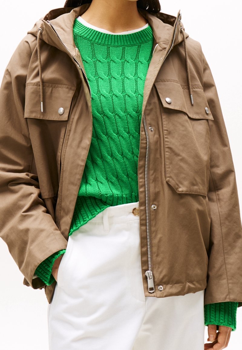 Bruine gematteerde capuchonjas met voorzakken, zilveren drukknopen en rits, gedragen over een groene kabelgebreide trui. Gecombineerd met een witte broek.