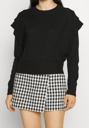Femme portant un pull noir à manches longues avec des épaules à volants et une mini-jupe à carreaux noir et blanc.
