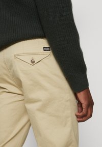 Dunkelgrüner Strickpullover mit gerippter Textur, kombiniert mit hellbeigen Hosen, die über eine dreieckige Gesäßtasche und ein Markenlabel verfügen.