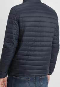 Marinblå puffjacka med en quiltad design, med horisontell söm, hög krage och elastiska ärmslut. Mjuk och lätt material.