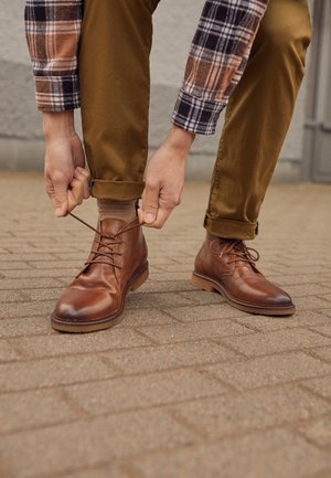 Persoon die veters strikt van bruine leren laarzen, draagt een bruine broek en een geruit overhemd, staat op een geplaveid oppervlak buiten.