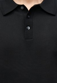 Polo noir en maille avec trois boutons noirs, col rabattur, porté par une personne.
