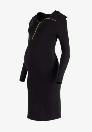 Zwarte geribde zwangerschapsjurk met een ritskraag, lange mouwen en een aangesloten silhouet dat ruimte biedt voor een groeiende buik.