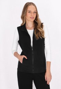 Gilet in pile nero con zip frontale, colletto alto e tasche laterali, indossato sopra una maglietta bianca a maniche lunghe. Texture liscia, design informale.