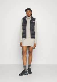 Gilet noir en duvet avec capuche, col roulé crème, jupe beige et chaussures de sport noires. Des chaussettes rayées et une casquette de baseball complètent le look.