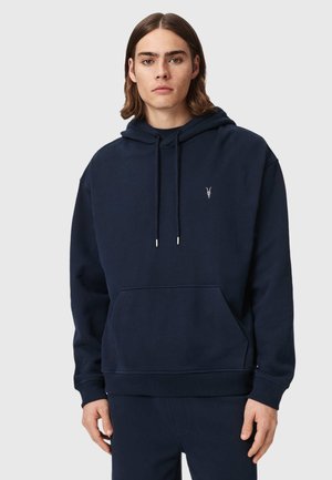 Jonge man met schouderlang haar die een donkerblauwe hoodie draagt met een voorzak en een klein wit geborduurd logo op de borst.