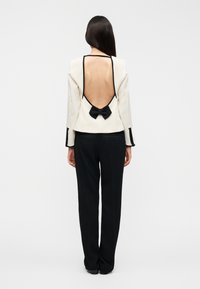 Πίσω όψη μιας εκρού ζακέτας με βαθιά πλάτη, μαύρη επένδυση και διακόσμηση φιόγκου, συνδυασμένη με μαύρο παντελόνι. Λείο υφασμάτινο υλικό.