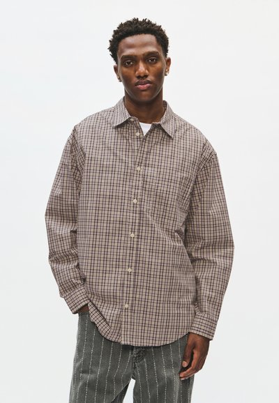 Jeune homme debout, les mains dans les poches, portant une chemise à carreaux beige et marron boutonnée et un pantalon sombre à rayures verticales blanches.