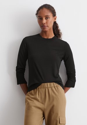 Chemise noire à manches longues avec un col rond et des poignets côtelés, associée à un pantalon beige à taille élastique avec une poche latérale.