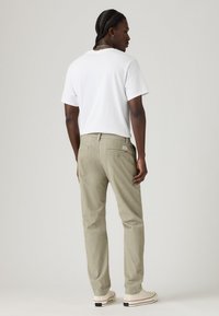 Man with braided hair wearing a white t-shirt, beige pants, and white sneakers, standing facing away against a plain background.