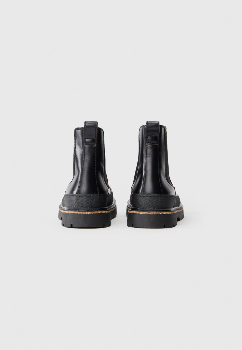 Une paire de bottines en cuir noir avec des languettes de traction, des semelles en caoutchouc épaisses et des coutures contrastantes, présentées de dos sur un fond blanc.