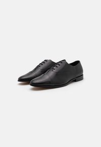Zapatos de vestir de piel negra con punta afilada, diseño con cordones, textura suave y detalle en la puntera de color contrastante. Suela plana de goma.