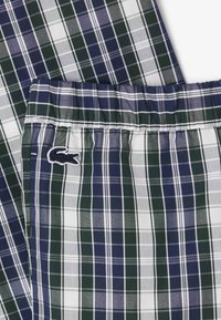 Pantaloni corti in cotone a quadri con strisce blu navy, verdi e bianche. Vita elastica con il logo del coccodrillo Lacoste ricamato di lato.