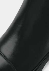 Gros plan d’une botte en cuir noir lisse et brillante avec des coutures visibles le long du bord de la semelle sur un fond clair.