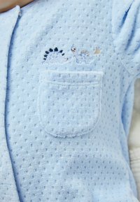 Mameluco de bebé de tela azul texturizada con mangas cortas, que presenta un pequeño bolsillo en el pecho y un diseño de dinosaurio bordado en colores claros.