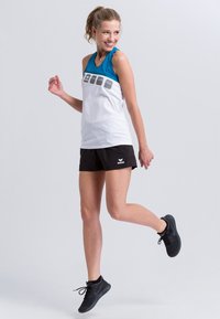 Canotta sportiva bianca con scollatura blu, decorata con un design grafico al centro. Abbinata a pantaloni corti neri e scarpe sportive nere.