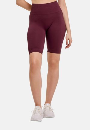 Vrouw draagt naadloze biker shorts met hoge taille in bordeaux en witte sportschoenen, staand voor een effen lichte achtergrond.