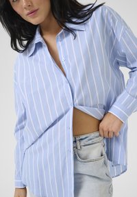 Cămașă butonată de un albastru deschis cu dungi verticale albe, cu mâneci lungi și o croială lejeră, parțial băgată în șorturi din denim cu talie înaltă.