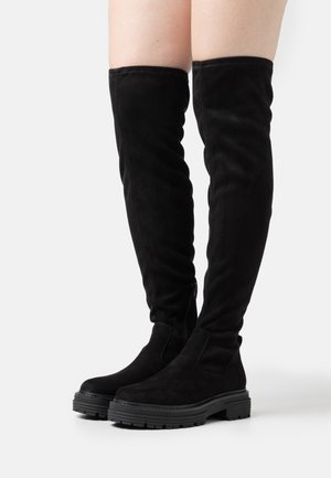 Bottes noires en daim au-dessus du genou avec semelles épaisses et texturées portées sur des jambes nues sur un fond blanc uni.