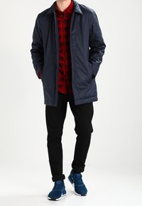Marineblaues, leichtes Jacket über einem roten Karoshirt, kombiniert mit schwarzen Hosen und blauen Sportschuhen. Verfügt über einen klassischen Kragen und einen Knopfverschluss.