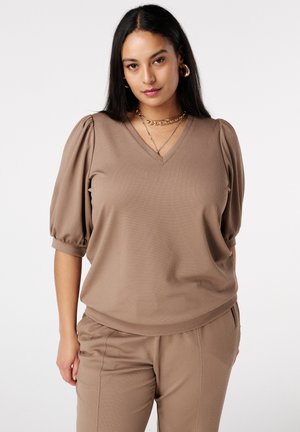 Vrouw draagt een taupekleurige top met pofmouwen en bijpassende broek, gecombineerd met gelaagde gouden kettingen en oorringen, poseert tegen een witte achtergrond.