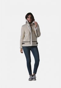 Veste en shearling blanche avec col montant, fermeture éclair frontale, deux poches latérales et accents en cuir noir. Associée à un jean bleu et des baskets.