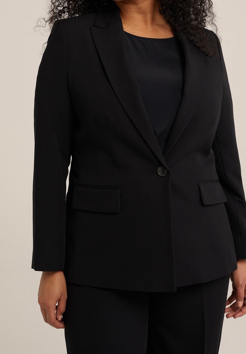 Femme portant un blazer noir sur mesure avec un seul bouton et des poches à rabat par-dessus un top noir, associé à un pantalon noir assorti.