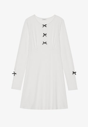 Biała sukienka z długim rękawem uszyta z prążkowanej tkaniny, z trzema czarnymi kokardami z przodu i dwiema na rękawach. Okrągły dekolt, w kształcie litery A.