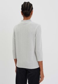 Pull en tricot gris clair avec un motif en grille texturé, manches trois-quarts et un petit col. Porté avec un pantalon sombre.