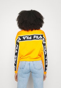 Felpa arancione con accenti neri del logo FILA sulle maniche e sul retro. Materiale morbido con una vestibilità rilassata, abbinata a jeans a vita alta.