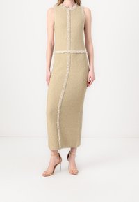 Robe sans manches beige texturée avec bord effiloché, longueur genou, présentant des coutures verticales et une silhouette ajustée. Sandales à talons transparents portées.