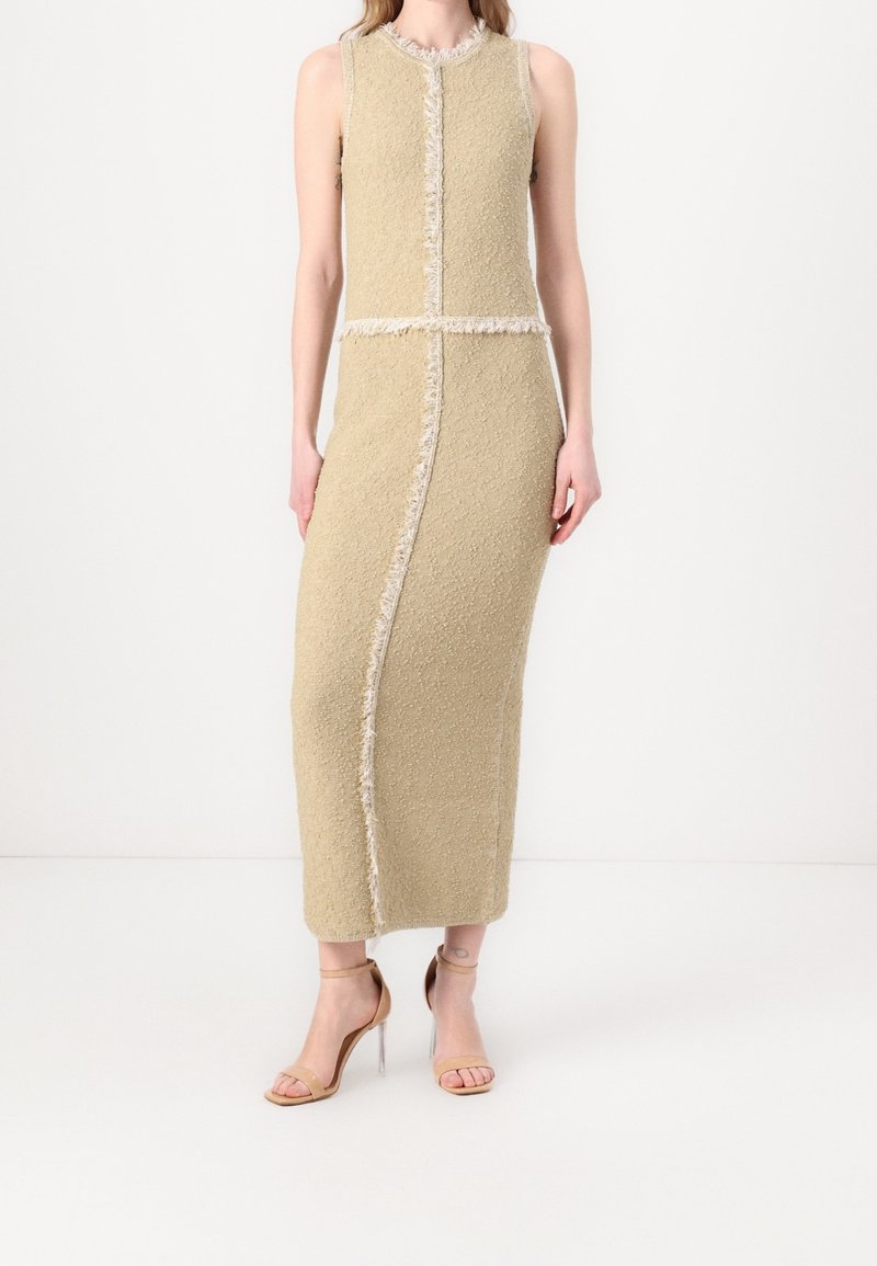 Robe sans manches beige texturée avec bord effiloché, longueur genou, présentant des coutures verticales et une silhouette ajustée. Sandales à talons transparents portées.