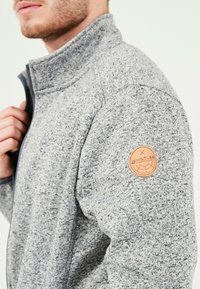 Grå fleecejacka med hög krage, försedd med en läderlogotyp på vänster axel och ett texturerat stickmönster.