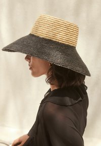 Bredbrättad hatt med en vävd design som har en beige ovansida och en svart undersida. Materialet verkar vara texturerat med en strukturerad form.