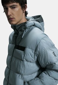 Gewatteerde hooded jas van lichtgrijs stof met een gladde textuur. Voorzien van een rits aan de voorkant, elastische manchetten en een contrasterend zwart zakdetail.