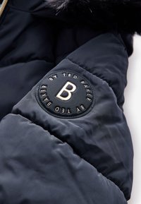 Mørkeblå pufferjakke med en glat, tekstureret overflade. Har et cirkulært logo-patch med "B" og "By Ted Baker". Guldhardware på lynlåsen.