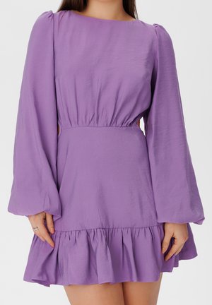 Mujer con un vestido mini morado de manga larga, cintura fruncida y dobladillo con volantes, de pie con las manos cerca de la falda.